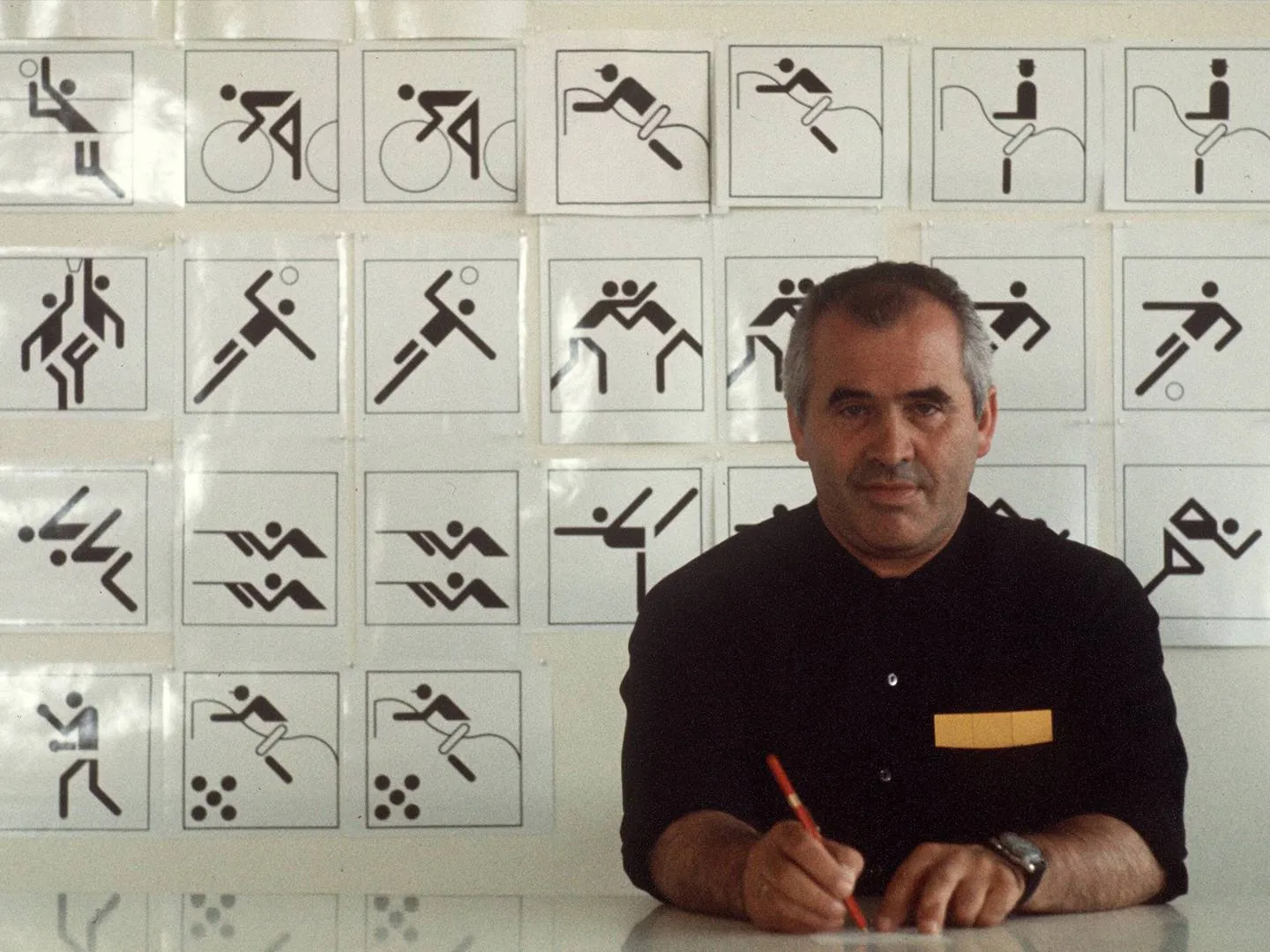 Otl Aicher sitzt vor Wand mit ikonischen Sportpiktogrammen. Sinnbild für klares Kommunikationsdesign, Markenentwicklung und visuelle Identität.